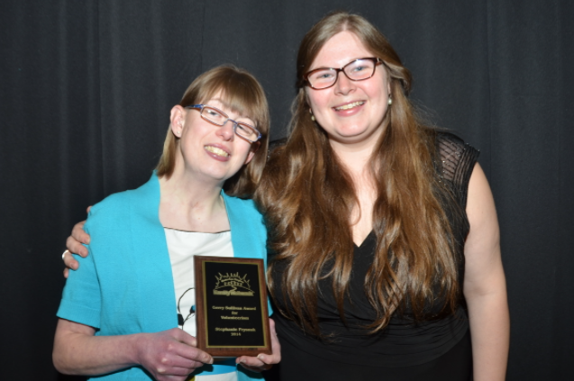 Stephanie Prysnuk holding up her award to the camera. She has short, light brown hair with bangs. She wears glasses and a bright blue cardigan over a white blouse. She is standing with her sister, who has her arm around Stephanie's shoulder. Both are smiling.